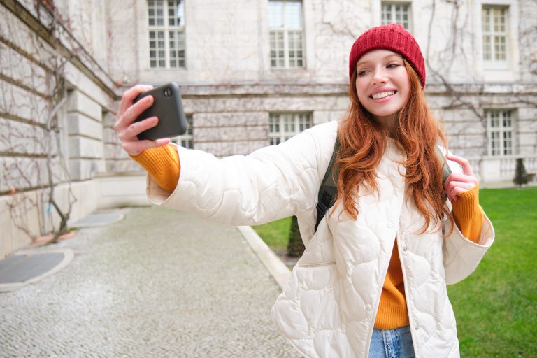 Portrait of happy girl tourist, takes selfie on smartphone in front of historical building, posing for photo on mobile phone camera